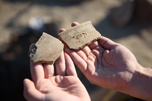 Two hands hold fragments of recently excavation pottery with a zig-zag design.