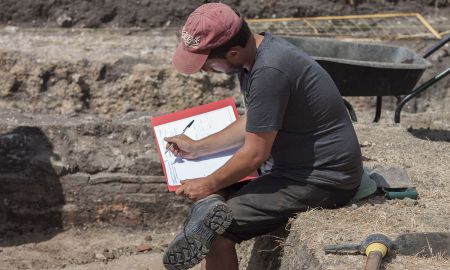 An archaeologist sits on the side of an excavation trench and completes context records.