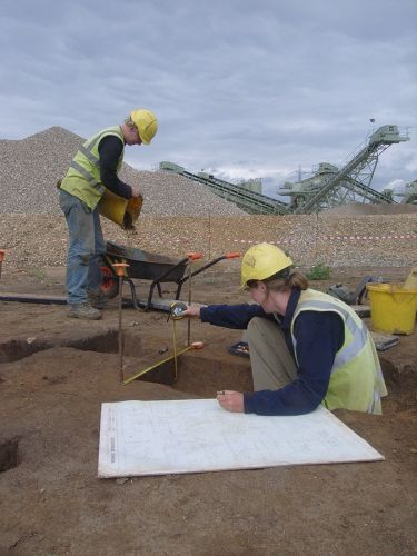 Two archaeologists excavate next to an industrial extraction site.