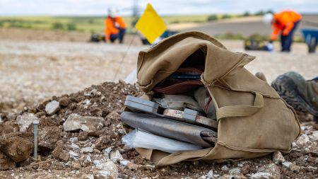A satchel containing a measuring tape and notebook lies on a dusty ground.