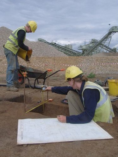 Two archaeologists excavate next to an industrial extraction site.