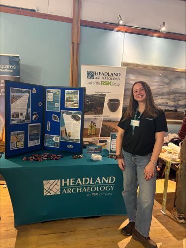 Photograph of Jess Wagstaff standing in front of a Headland Archaeology display stand