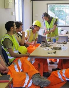 Archaeologists in hi-vis clothing sit around a table to examine finds with a specialist.
