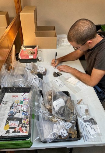 An archaeologist surrounded by finds writes on clear plastic finds bags at a desk.