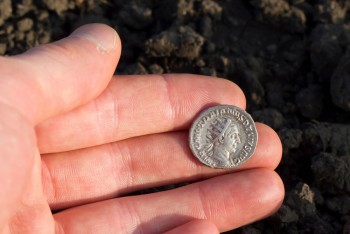 A Roman coin in the hand of an archaeologist, with disturbed earth in the background.