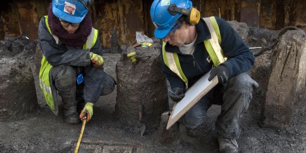 Two archaeologists in blue hard hats examine an archaeological feature.