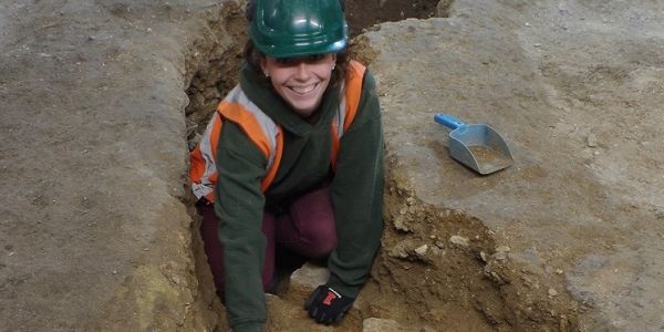 A female archaeologist, in high-vis jacket and hard hat, cleaning a trench.