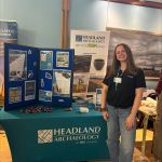 Photograph of Jess Wagstaff standing in front of a Headland Archaeology display stand